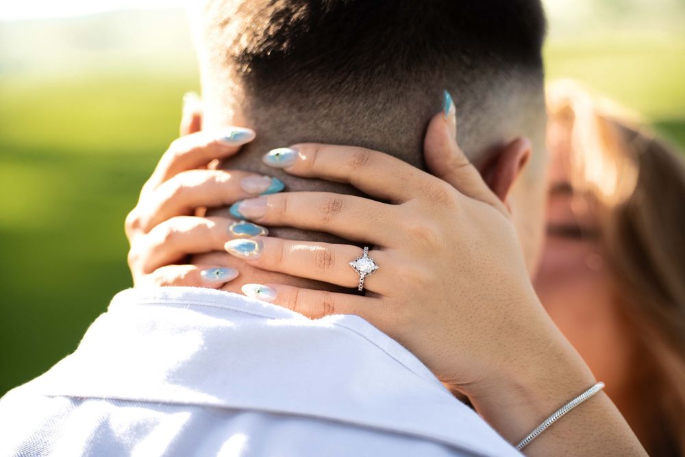 Close-up of a woman's hand with blue nails and a diamond engagement ring resting on a man's neck.