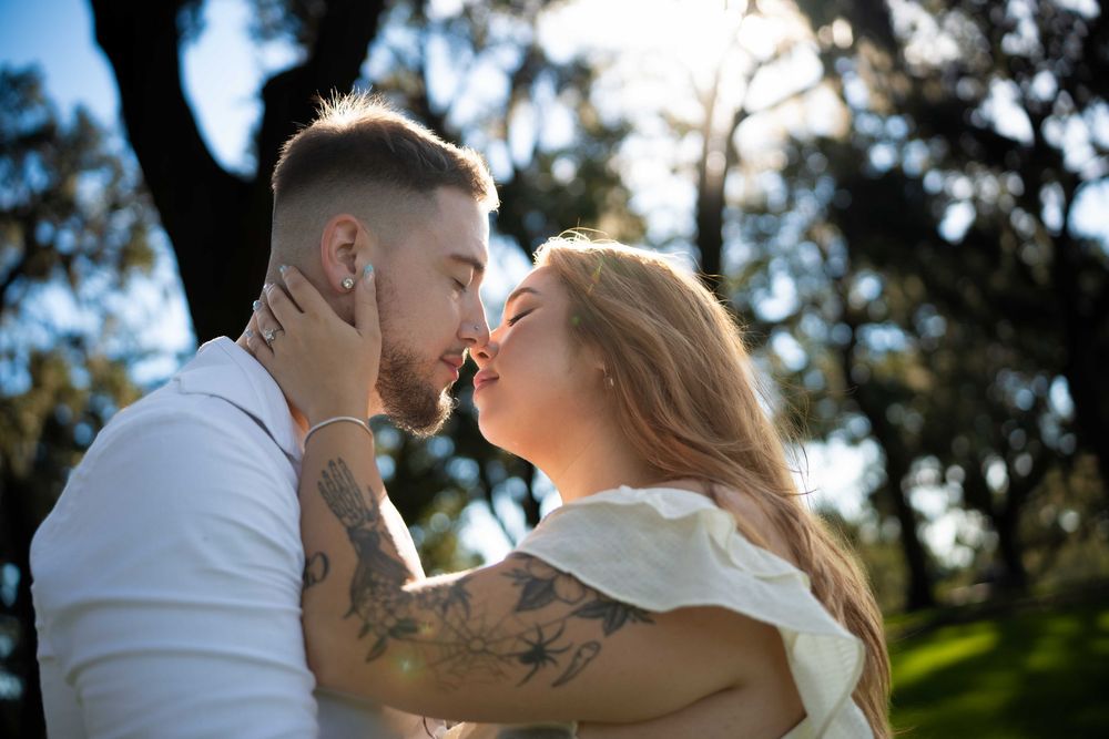 Couple sharing a romantic kiss outdoors, woman with tattoos embracing man in sunlit park setting.