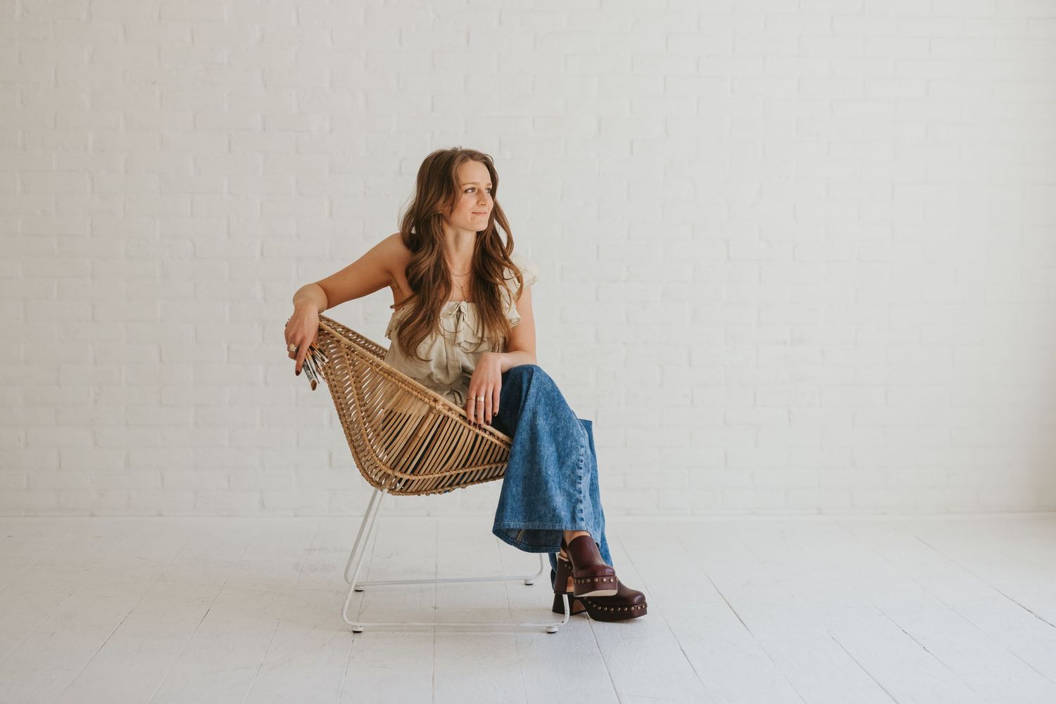 Woman in floral top and jeans sits in wicker chair against white brick wall, posing with crossed legs.
