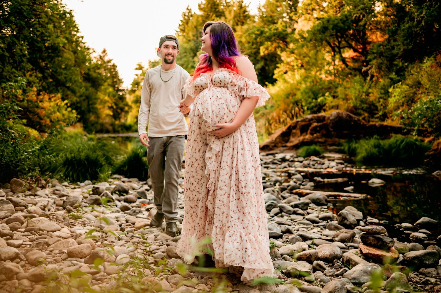 Couple poses in sunlit forest, pregnant woman in floral gown cradling baby bump during maternity photo session.