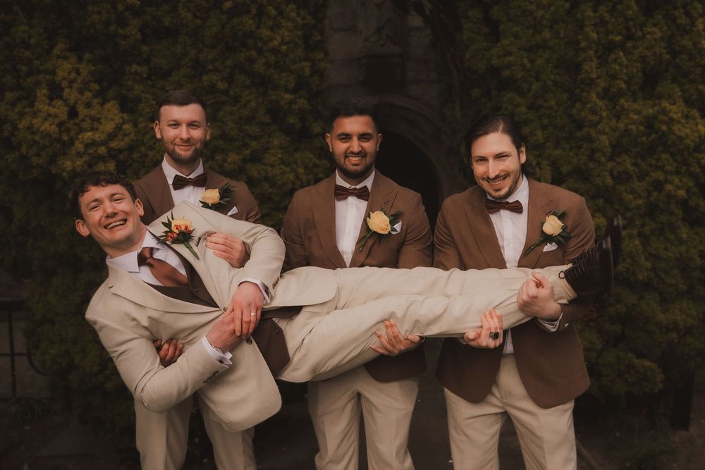 Groomsmen in brown suits with bow ties playfully carry the groom in a beige suit outdoors at a wedding.
