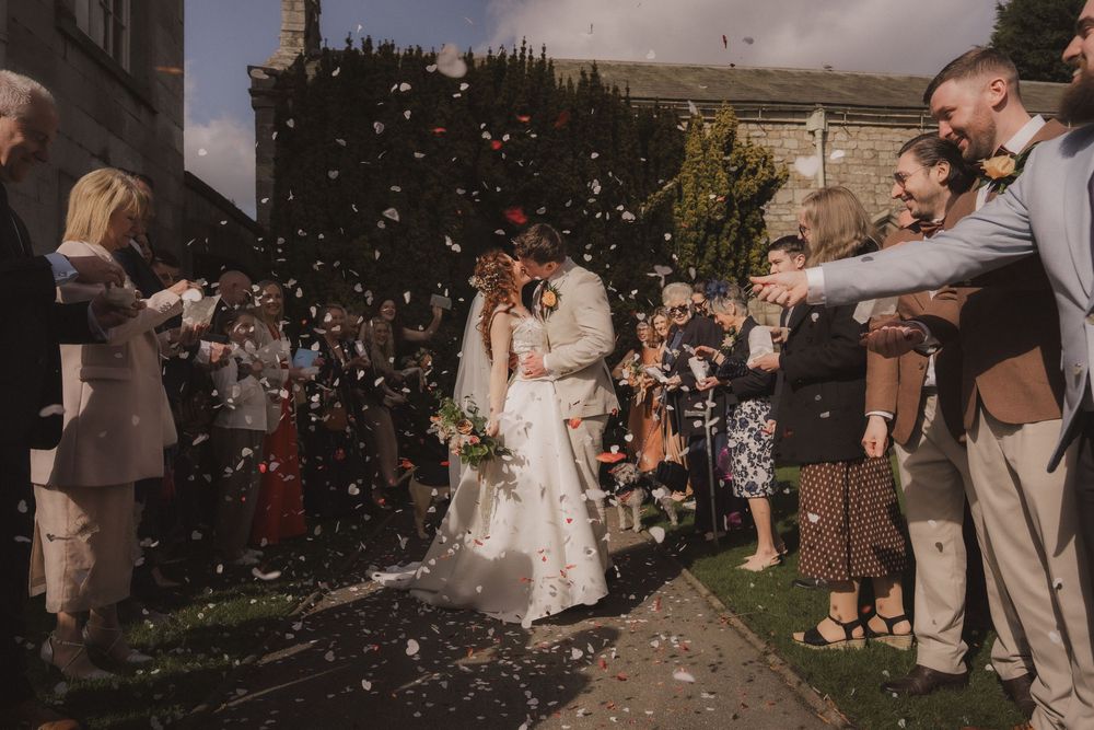 Bride and groom kissing amid confetti shower surrounded by cheering wedding guests outside a stone church.