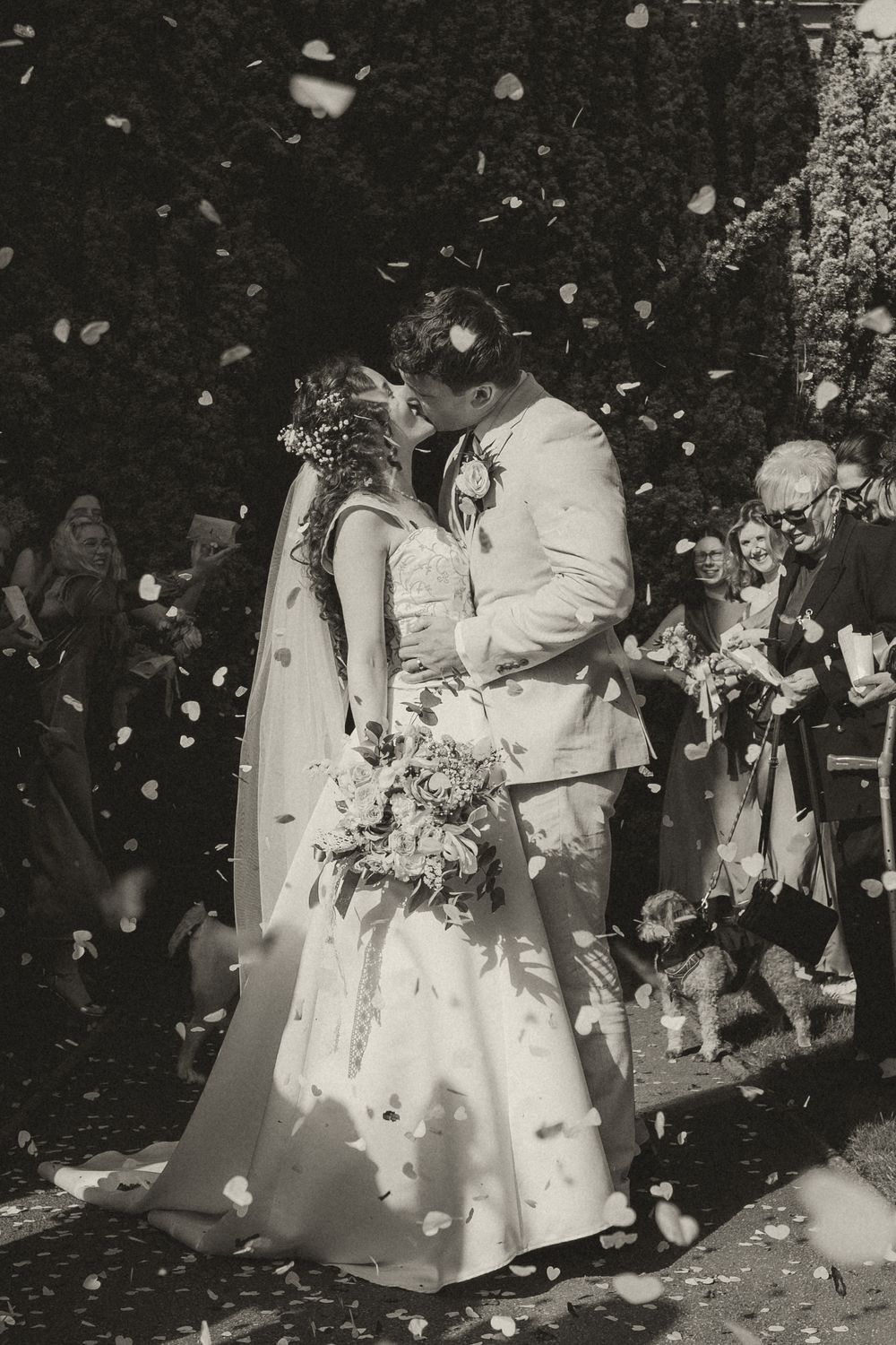 Black and white photo of bride and groom sharing first kiss surrounded by falling confetti petals with wedding guests.