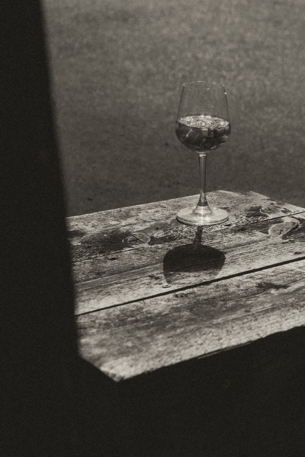 Black and white photo of a wine glass casting a shadow on a rustic wooden table in dramatic lighting.
