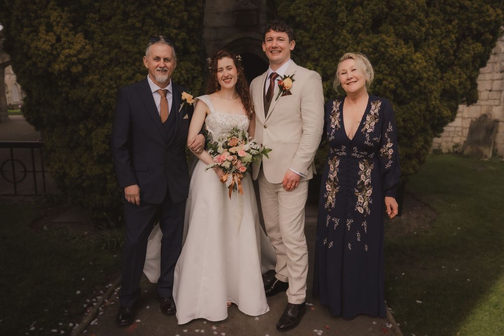 Wedding party of four posing outdoors, bride in white gown with bouquet, groom in beige suit, flanked by family.