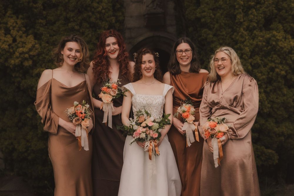 Bride poses with four bridesmaids in satin earth-tone dresses holding colorful bouquets outdoors.