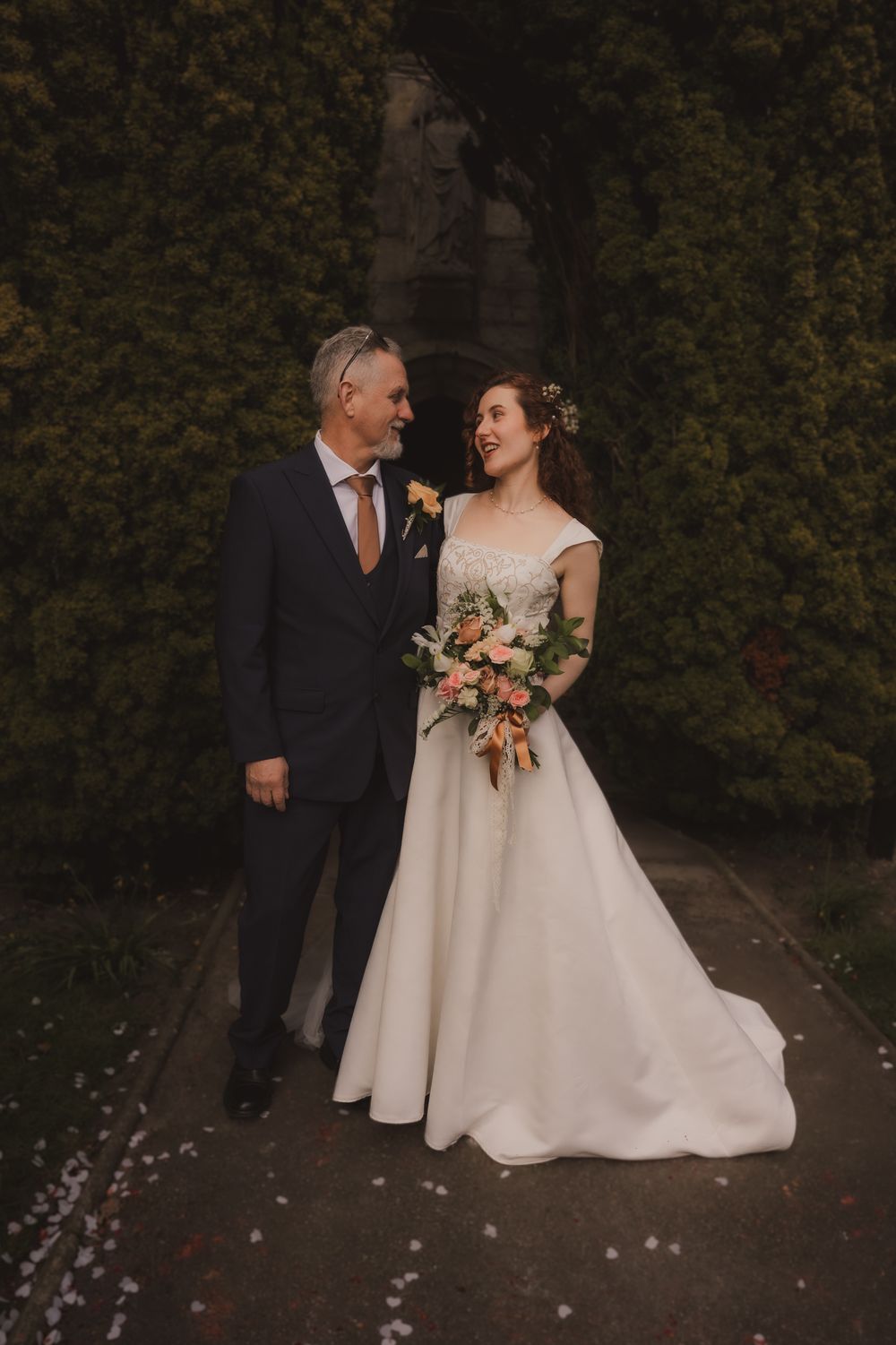 A bride and groom share a romantic moment outdoors, she holds a peach bouquet in a white ballgown.