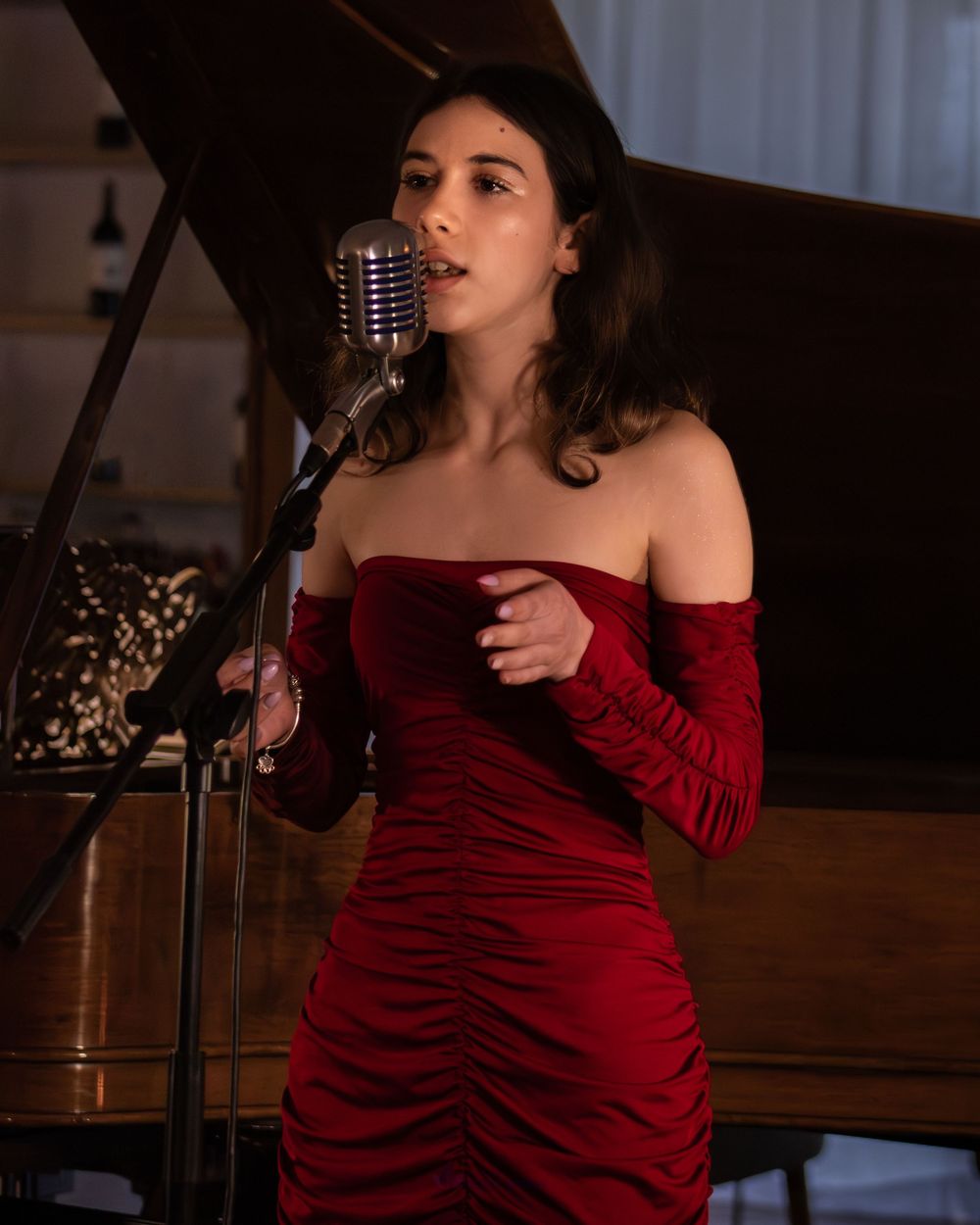 A woman in a red off-shoulder ruched dress sings into a vintage microphone beside a grand piano.