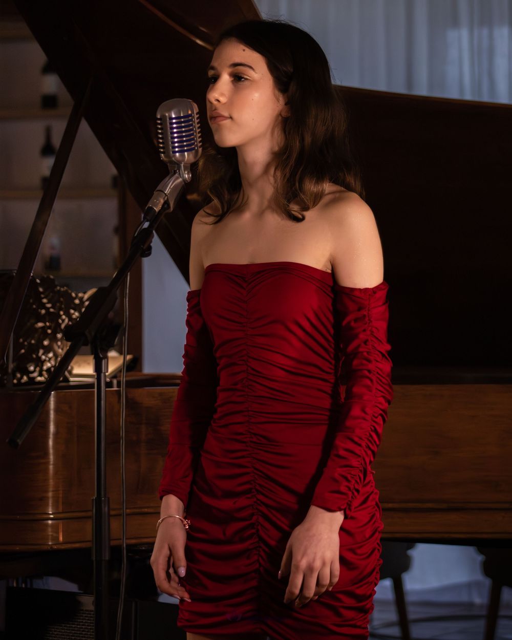 Young woman in red off-shoulder ruched dress singing at vintage microphone beside a grand piano.