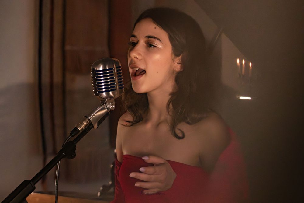 Young woman in red dress singing passionately into a vintage microphone on stage in a dimly lit venue.