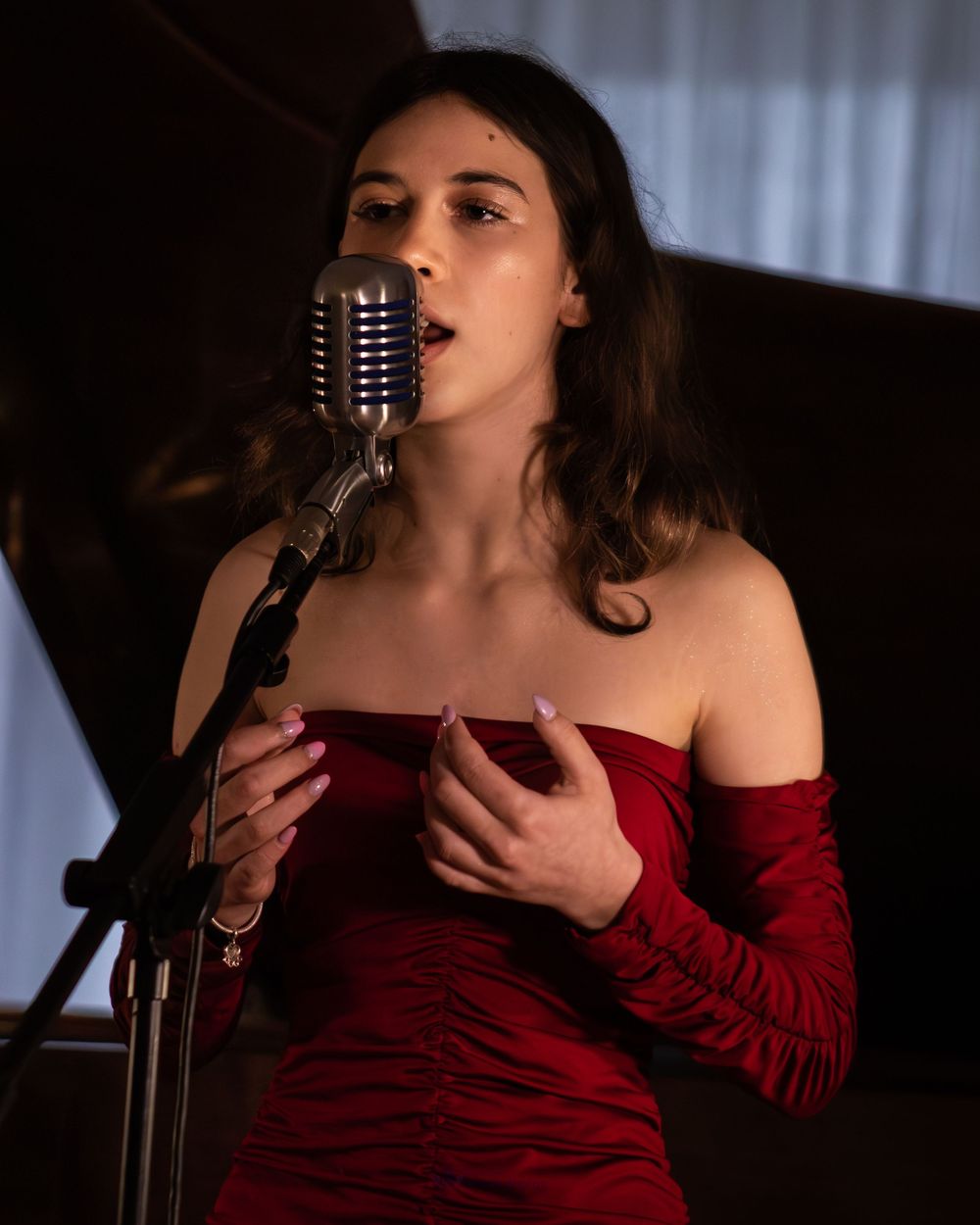 Woman in red off-shoulder dress singing passionately into a vintage microphone on stage.