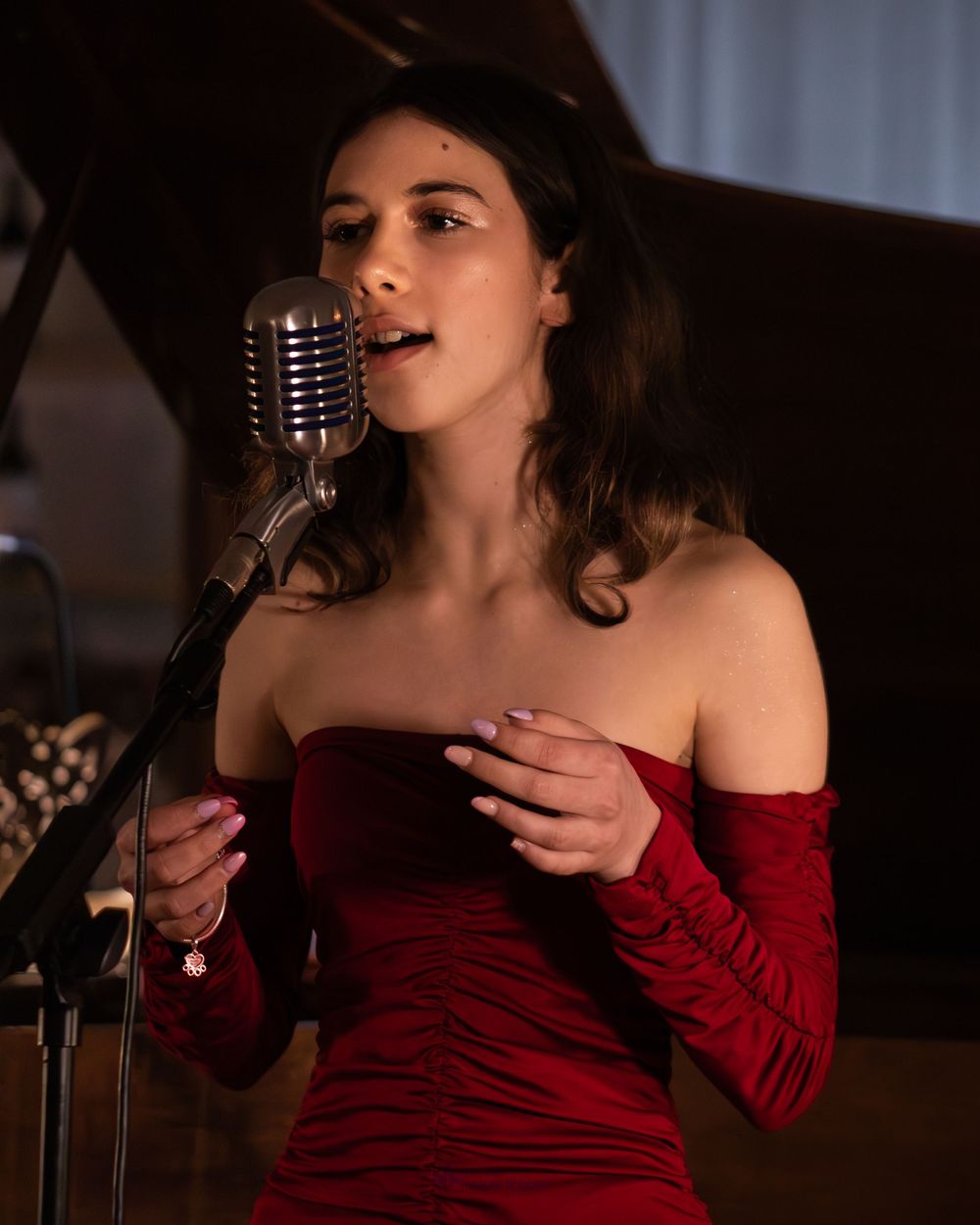 Young woman in red off-shoulder dress singing into vintage microphone at elegant musical performance.