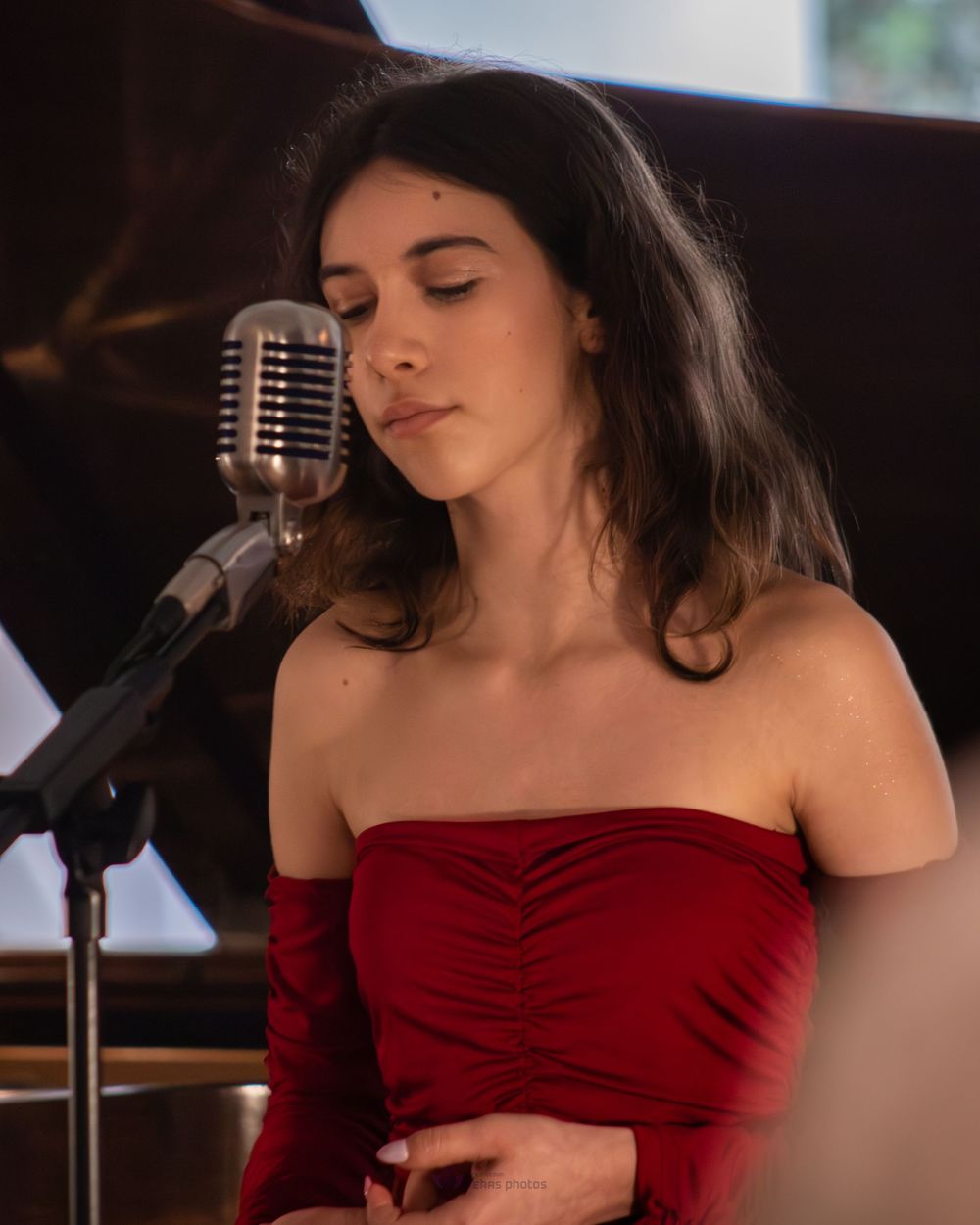 Young woman in red strapless dress singing into vintage microphone with eyes closed on stage.