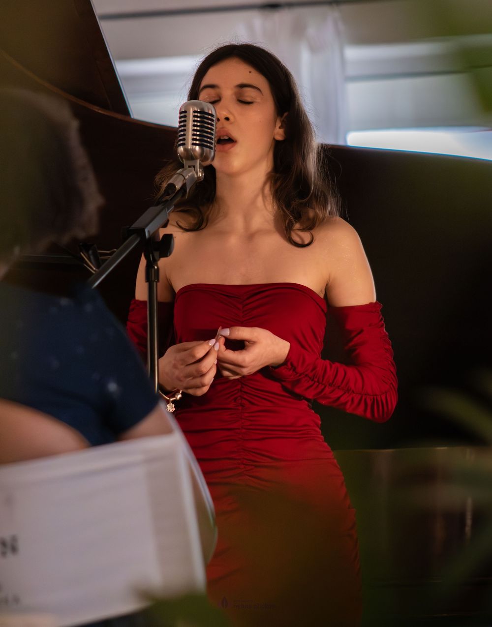 Woman in red off-shoulder dress singing into vintage microphone at an intimate indoor performance.