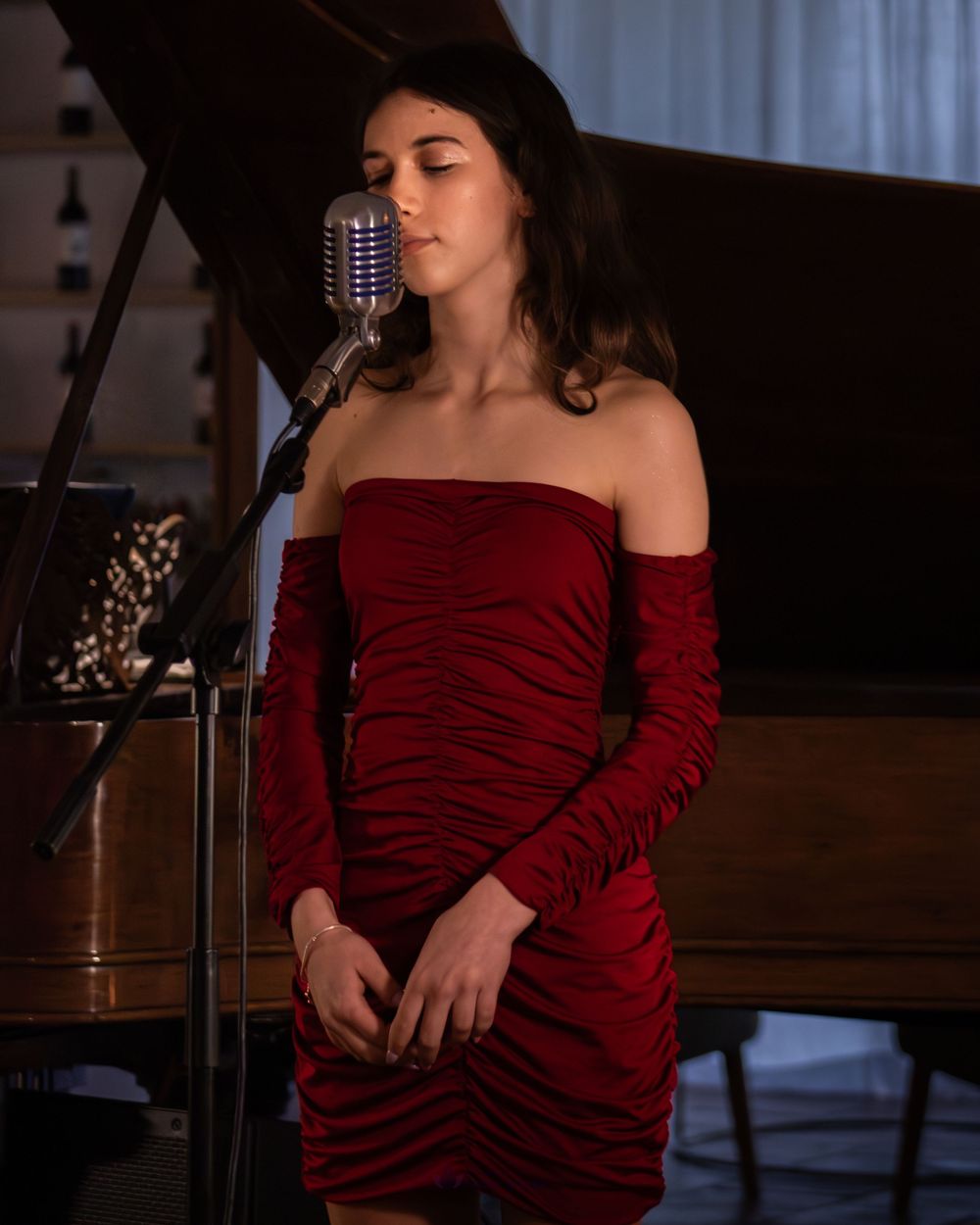 Young woman in red off-shoulder dress singing into vintage microphone near grand piano.