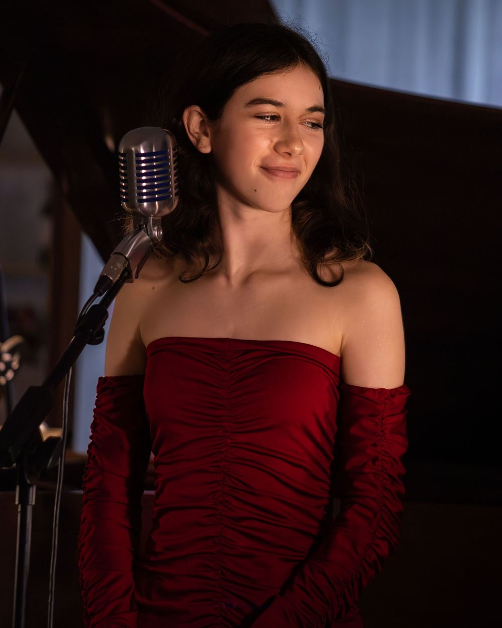 Young woman in red off-shoulder dress smiling near vintage microphone on stage.