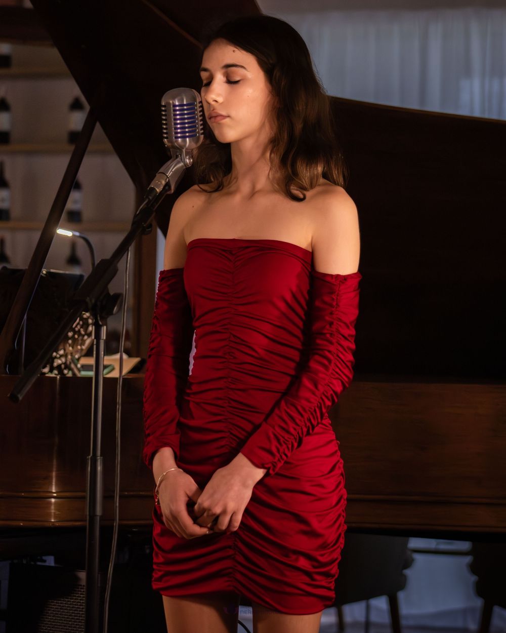 Young woman in red ruched off-shoulder mini dress singing at vintage microphone near a piano.