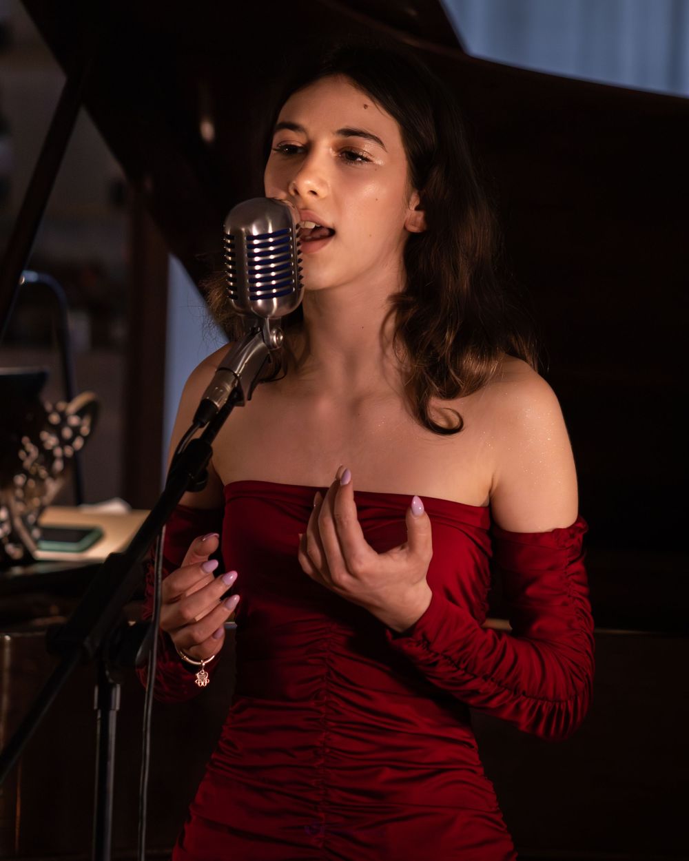 Young woman in red off-shoulder dress singing into vintage microphone at elegant indoor performance.