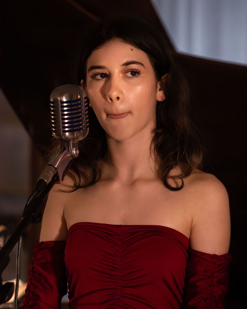 Young woman in red off-shoulder dress singing at a vintage silver microphone on stage.
