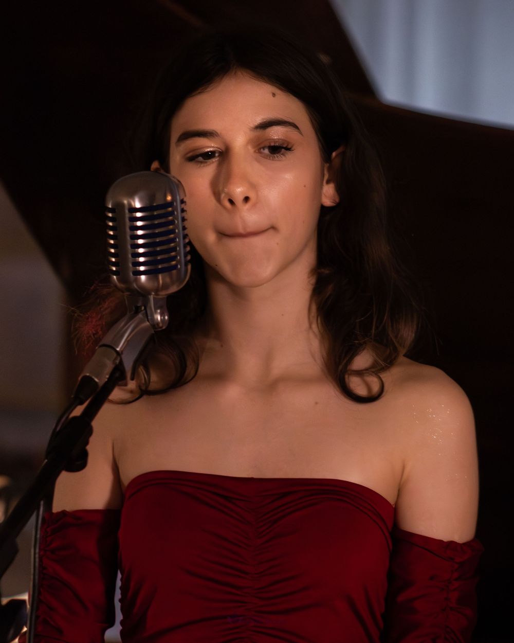 Young woman in red off-shoulder dress singing into a vintage silver microphone at a formal event.