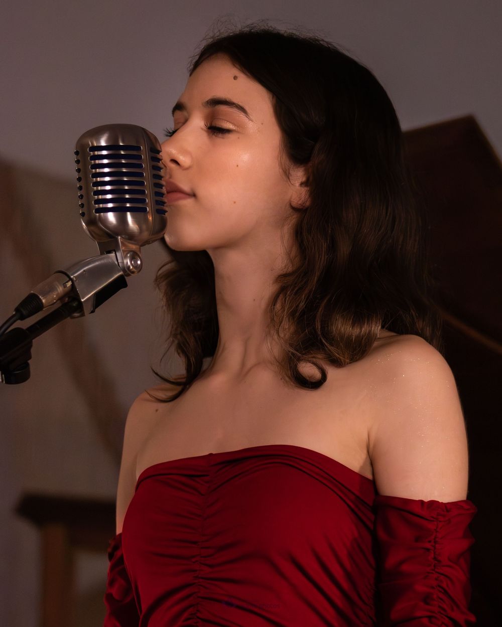 Young woman in red off-shoulder dress singing into a vintage silver microphone with eyes closed.