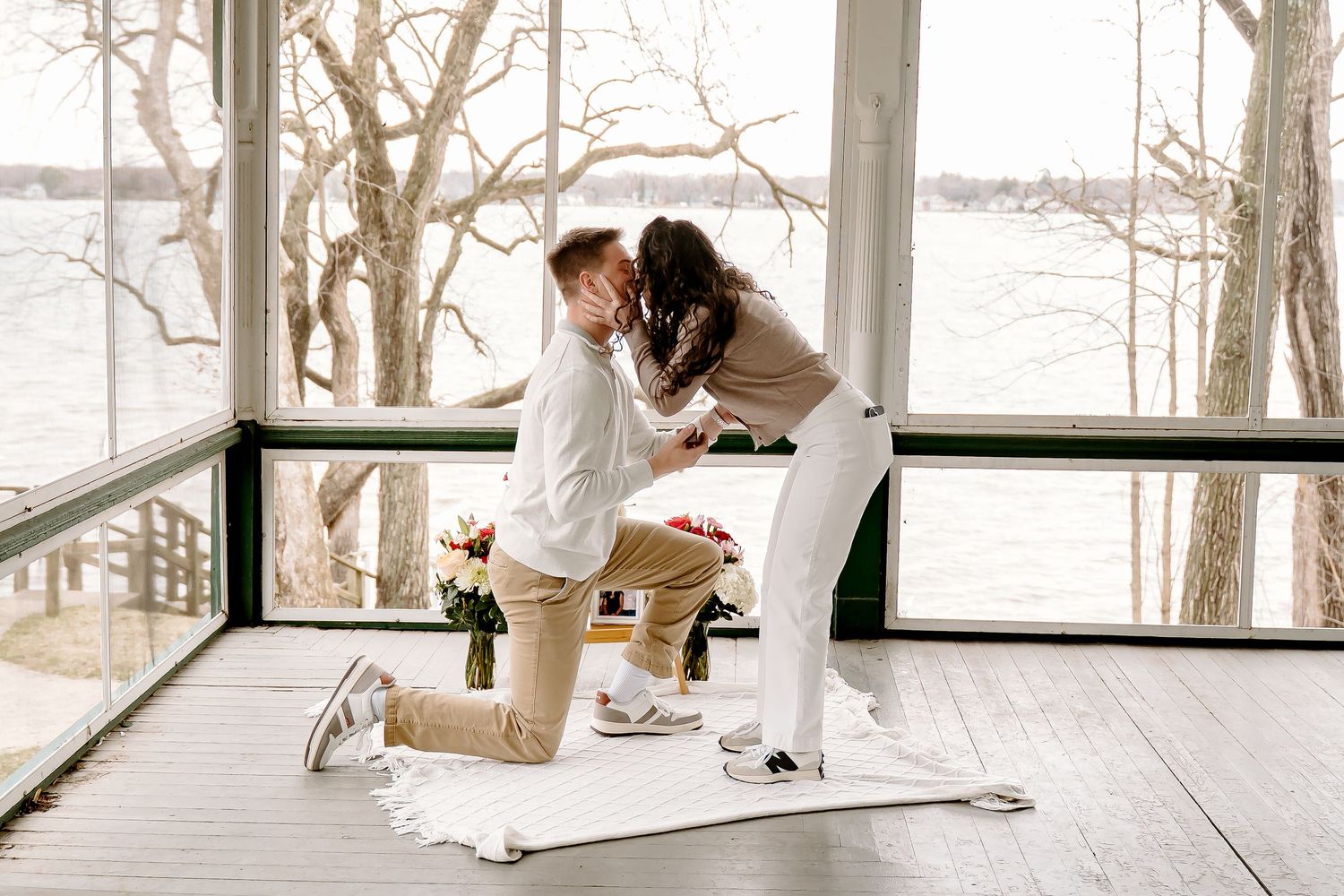 Man proposes on one knee to his partner in a sunlit lakeside cabin with flowers and a white blanket.
