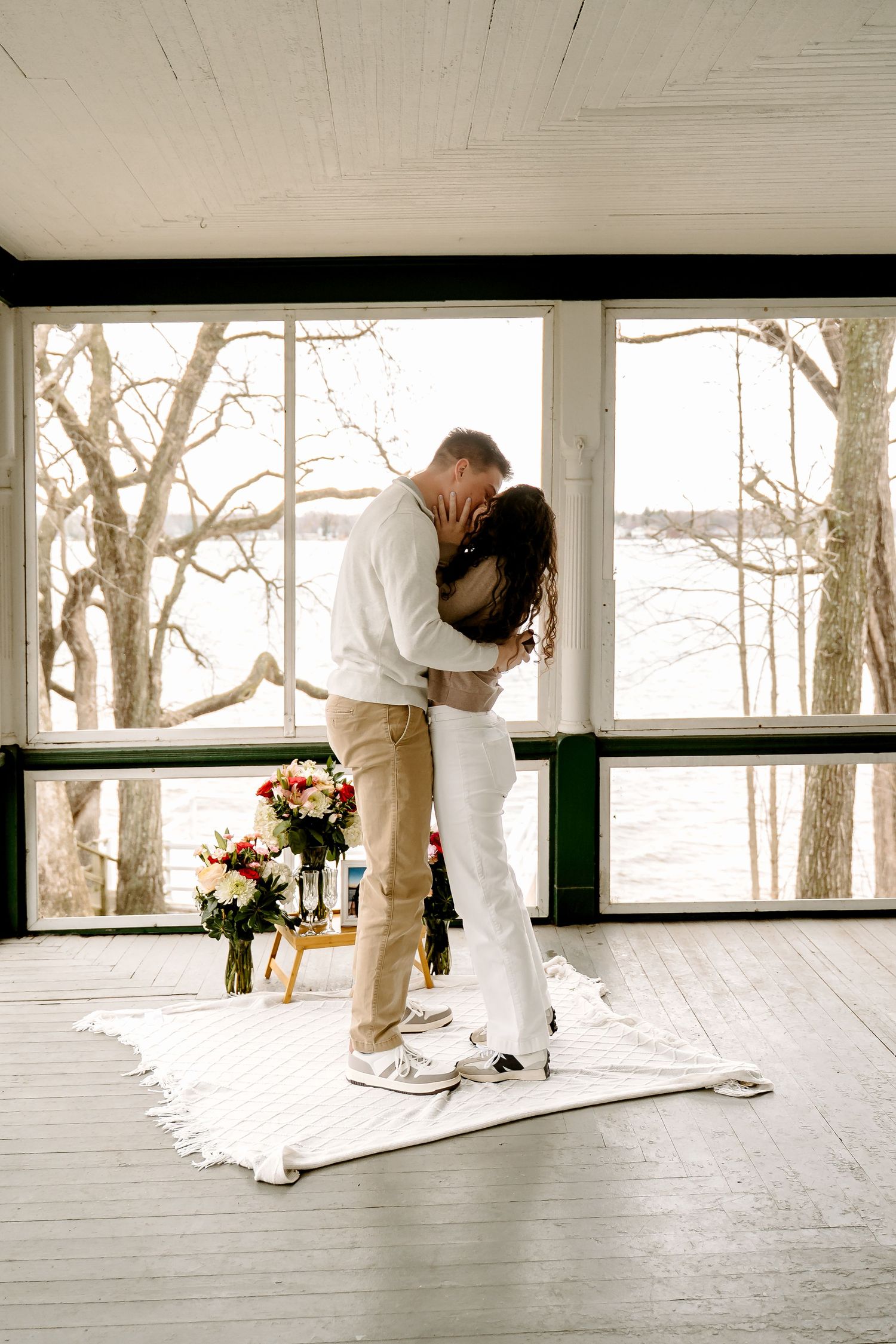 Couple sharing first kiss in white wedding attire on a blanket indoors with floral bouquet and snowy trees outside.