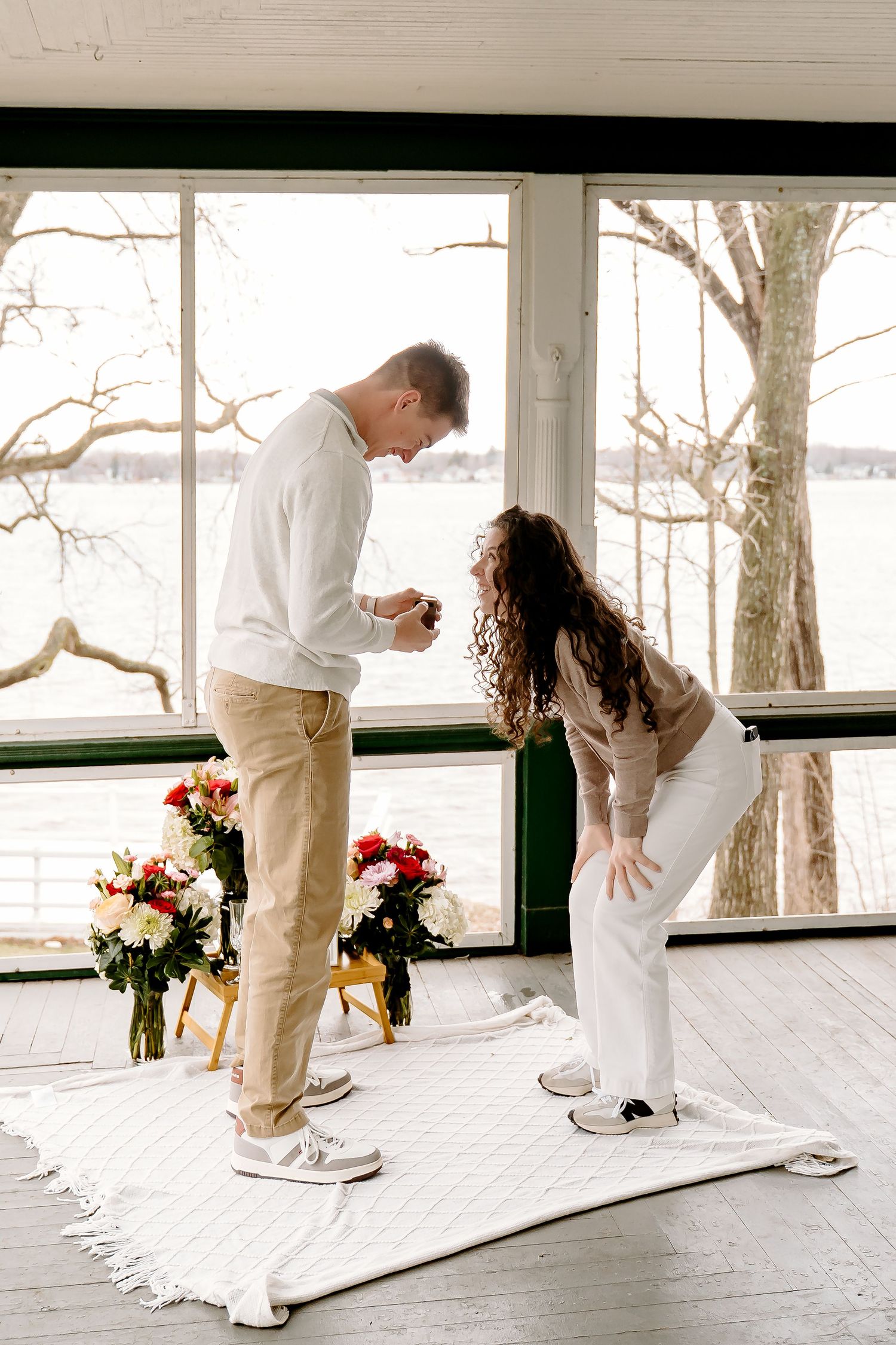 Man proposing to woman on white rug with floral arrangements, large windows overlooking winter landscape.