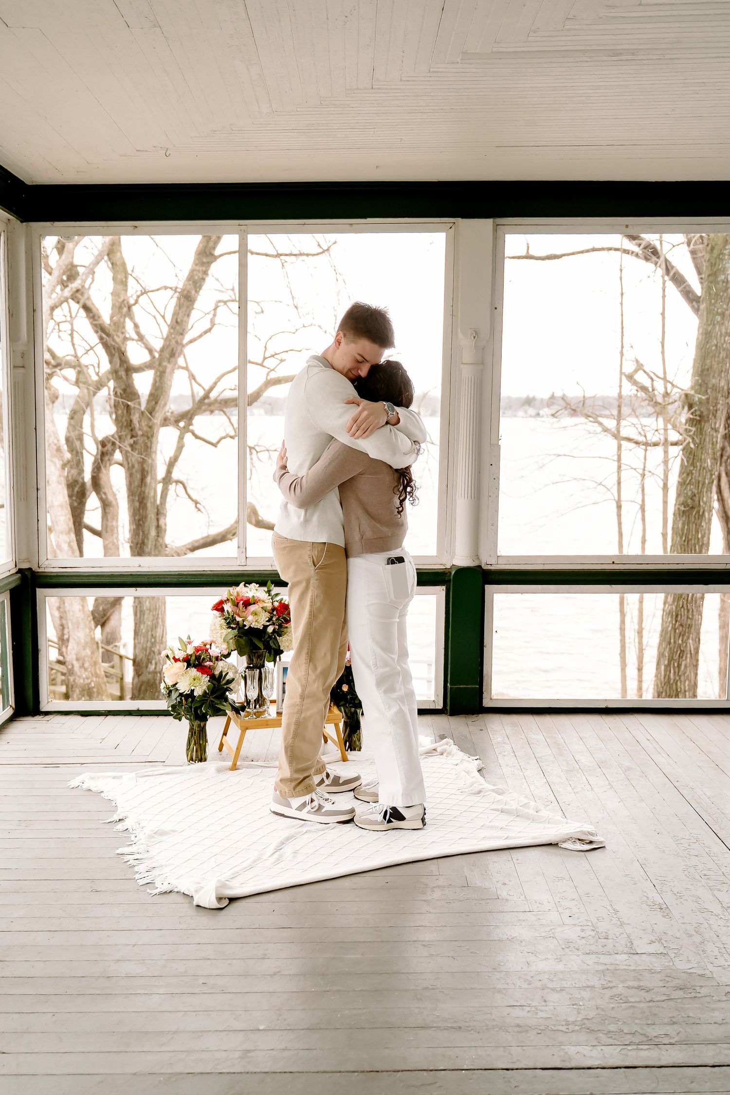 Couple sharing a romantic embrace on a white rug in a sunlit porch with floral arrangement nearby.