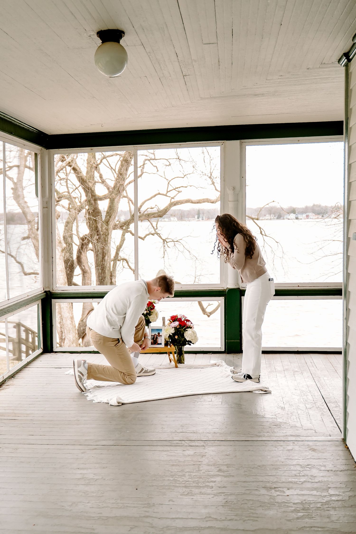 Man proposing on one knee to woman on white rug with red roses on sunlit porch overlooking lake.