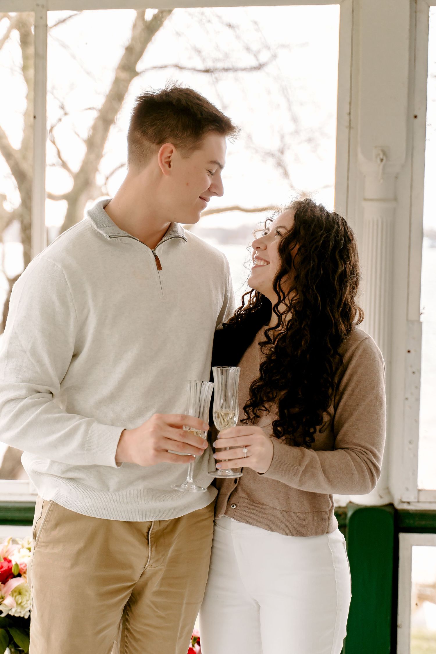 Couple toasting with champagne flutes indoors, smiling at each other, man in gray pullover and woman in tan cardigan.