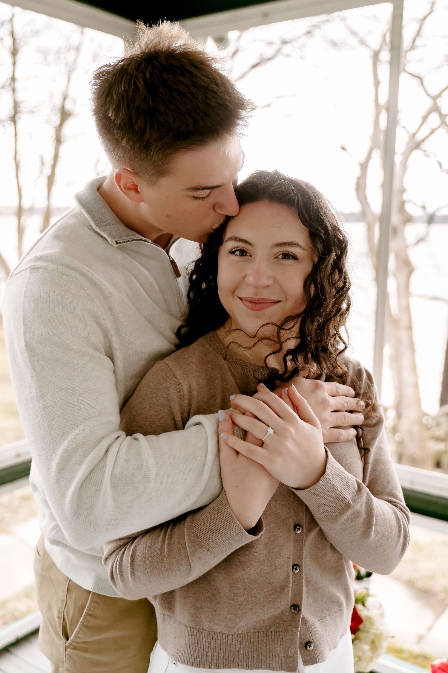 Couple embracing in a gazebo, man kissing woman's forehead while she smiles, wearing neutral tones.