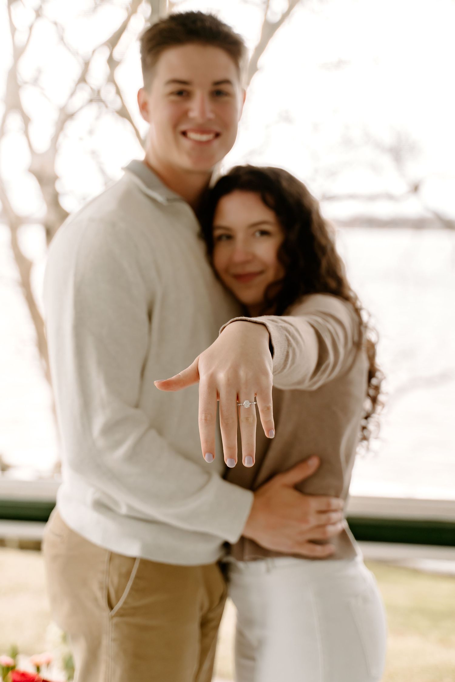 Happy couple showing off engagement ring by waterfront, woman extending hand with diamond ring while man embraces her.