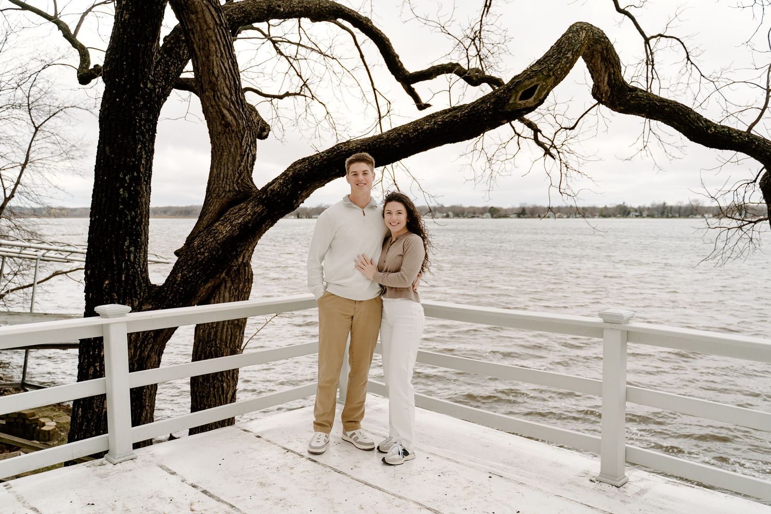 Couple embracing on a snowy lakeside dock under a large bare tree in winter casual attire.
