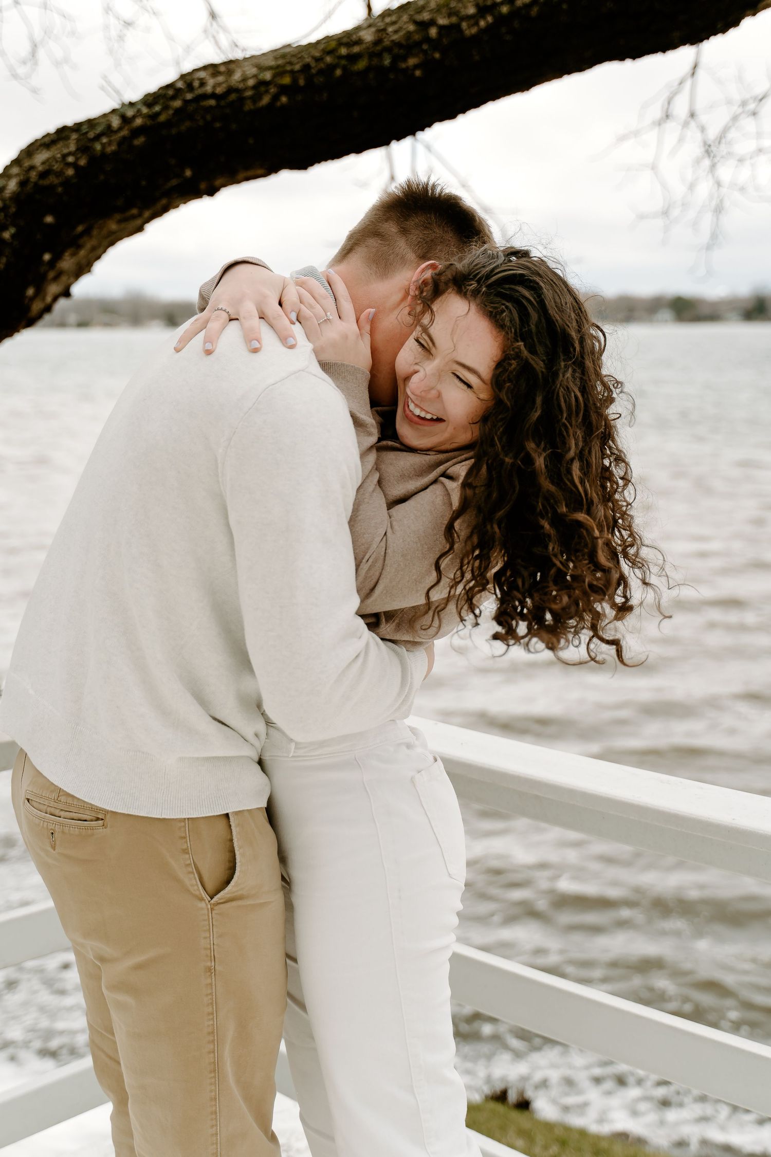 Joyful couple laughing and embracing on waterfront dock, woman with curly hair smiling as man nuzzles her neck.
