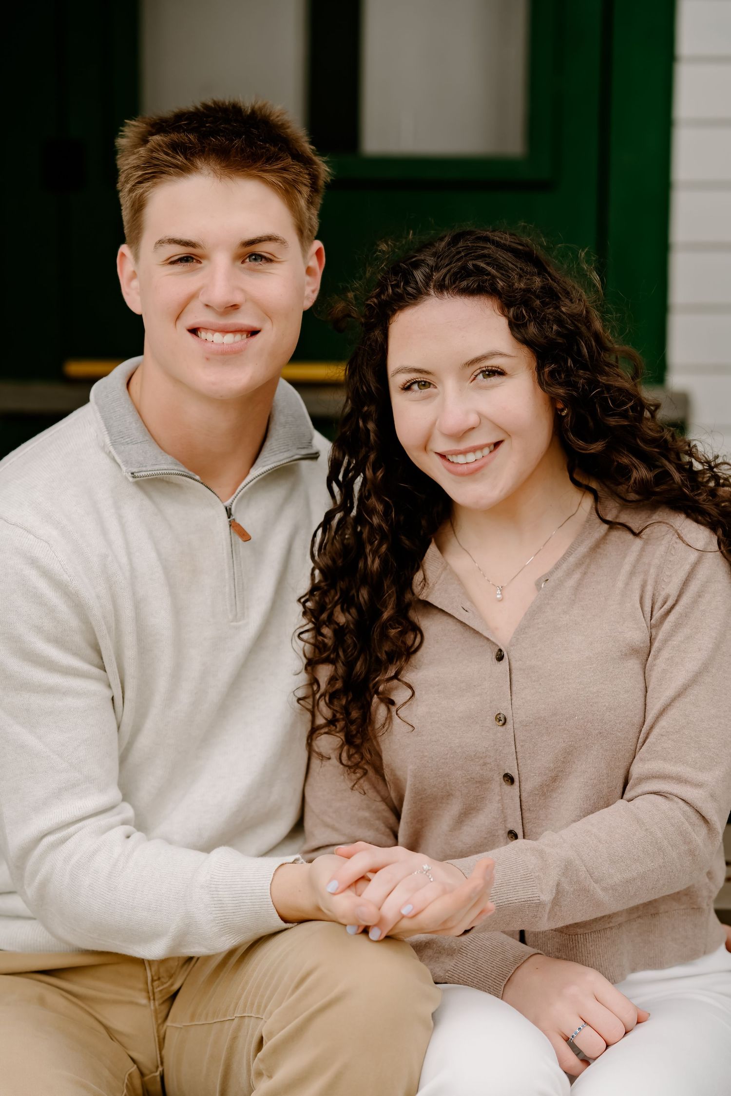 A smiling young couple in neutral tones sitting together holding hands in front of a green door.