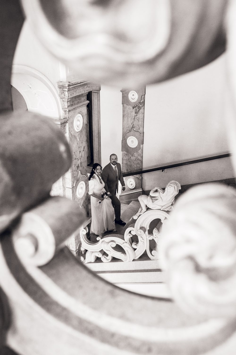 Black and white wedding photo of couple standing on grand staircase viewed through ornate architectural details.