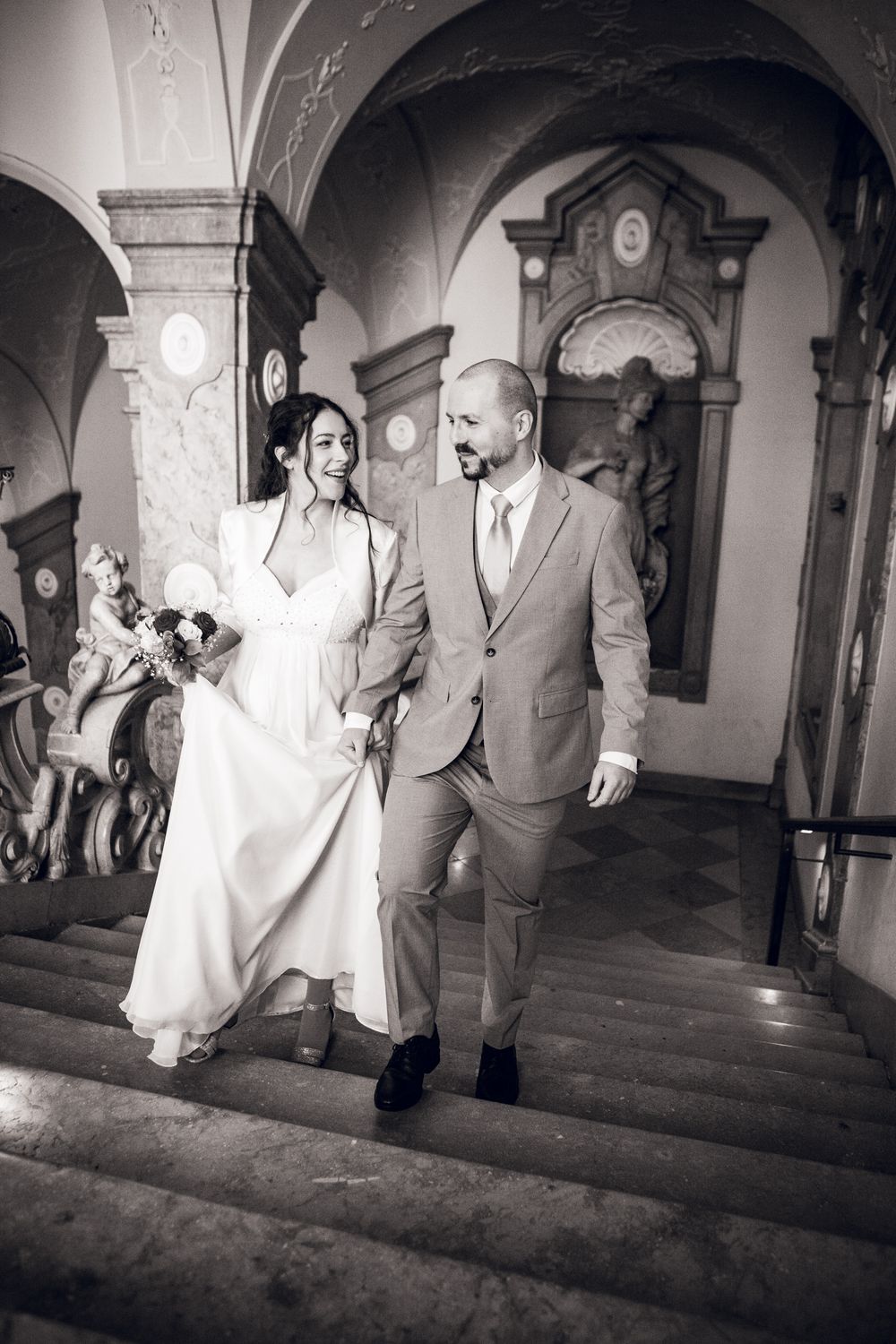 Black and white photo of a smiling bride and groom walking down elegant stairs in a historic building.