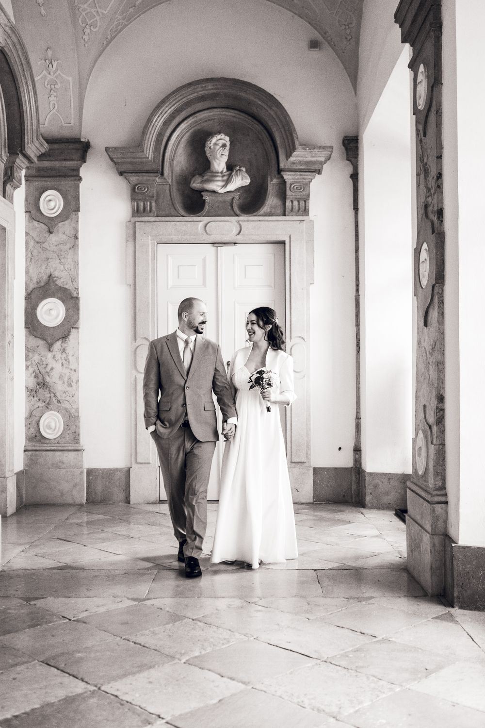Elegant black and white wedding photo of bride and groom walking through ornate marble hallway with classical architecture.