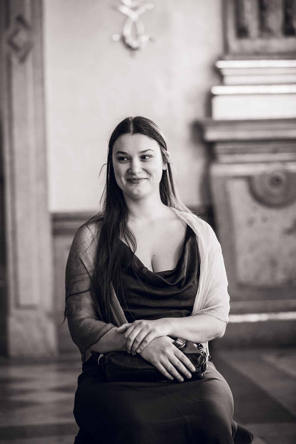 Black and white portrait of a young woman with long hair sitting gracefully in an ornate classical interior.