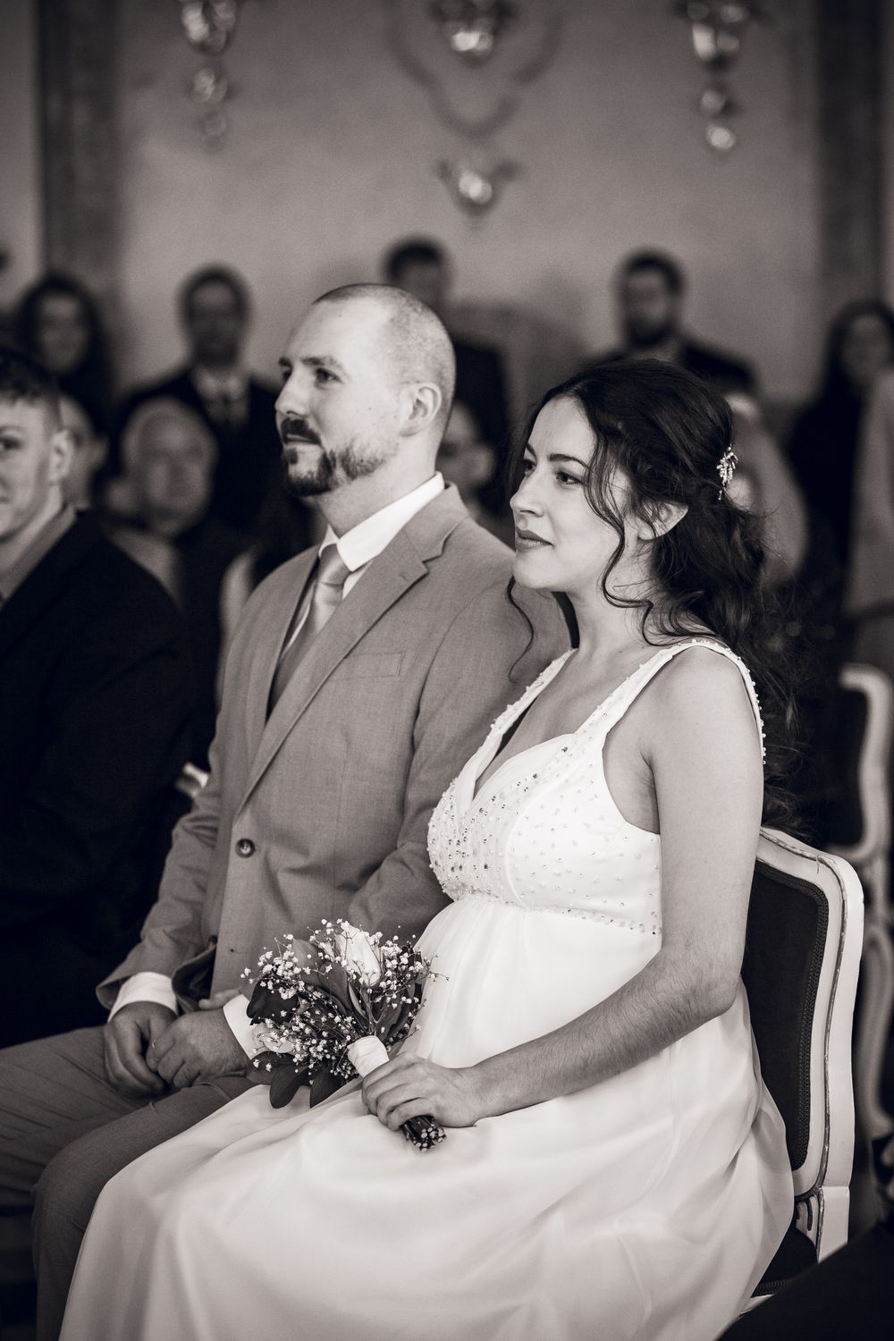 Black and white wedding photo of bride holding bouquet and groom seated during ceremony.