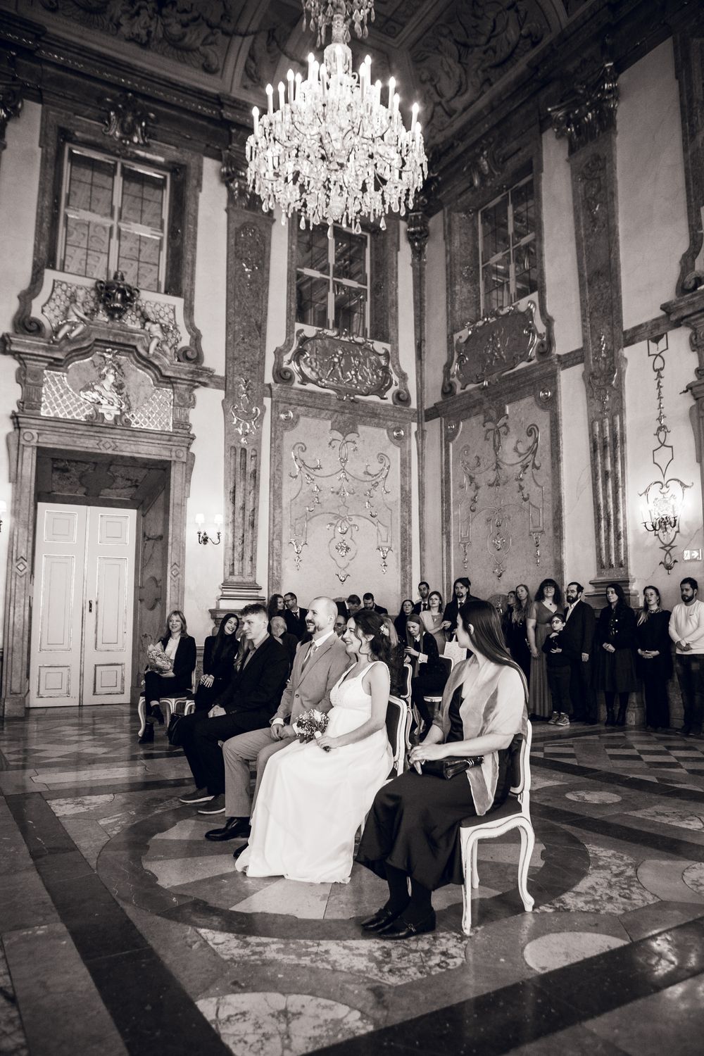 Black and white photo of a wedding ceremony in an ornate baroque hall with chandelier and seated guests.