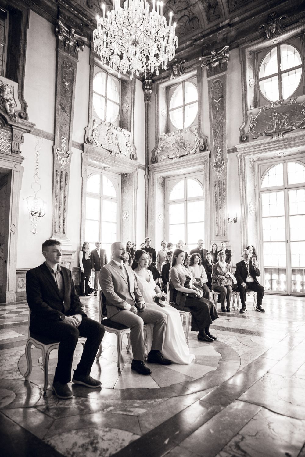 Black and white photo of wedding guests seated in an elegant baroque ballroom with chandelier during ceremony.
