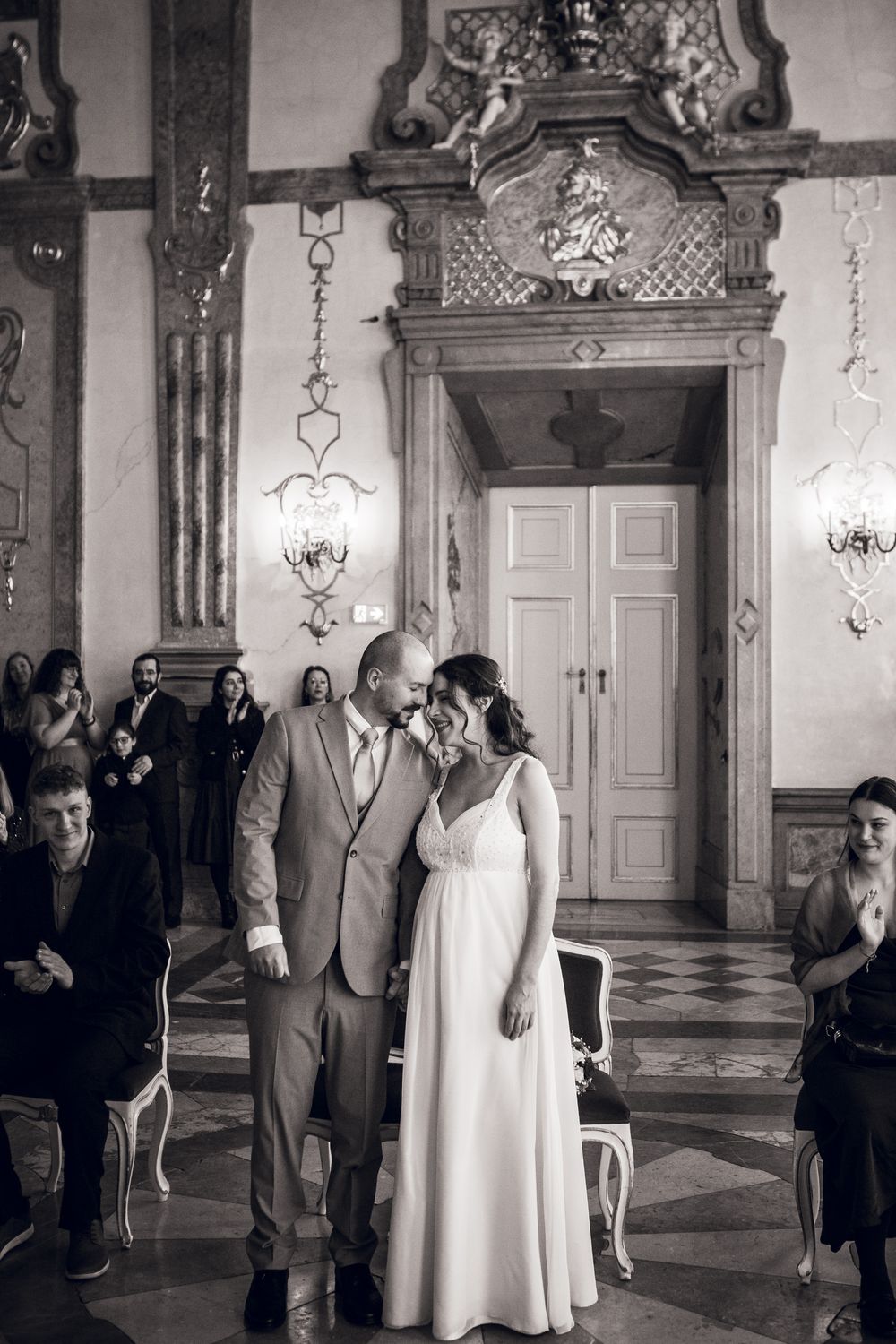 Black and white photo of bride and groom sharing an intimate moment during wedding ceremony in ornate historic venue.