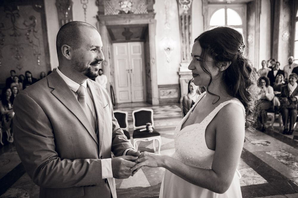 Bride and groom exchanging vows and rings in a grand hall, smiling joyfully in black and white photo.