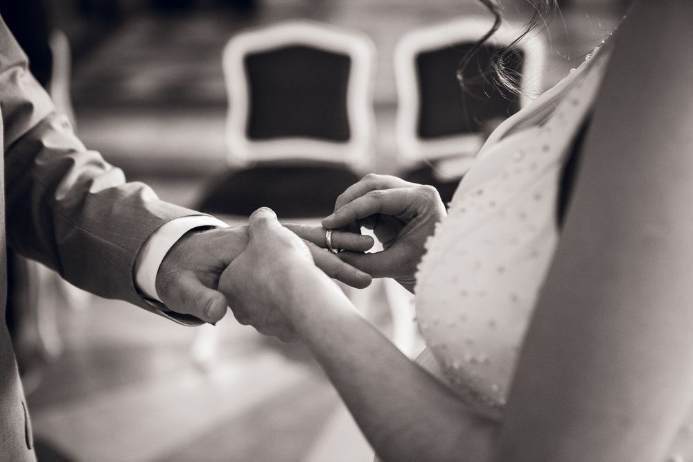 Black and white photo of bride placing wedding ring on groom's finger during marriage ceremony.