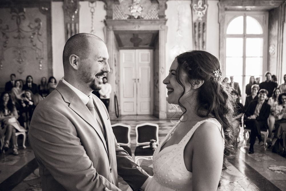 Black and white wedding ceremony photo of smiling bride and groom holding hands at altar with guests seated behind.
