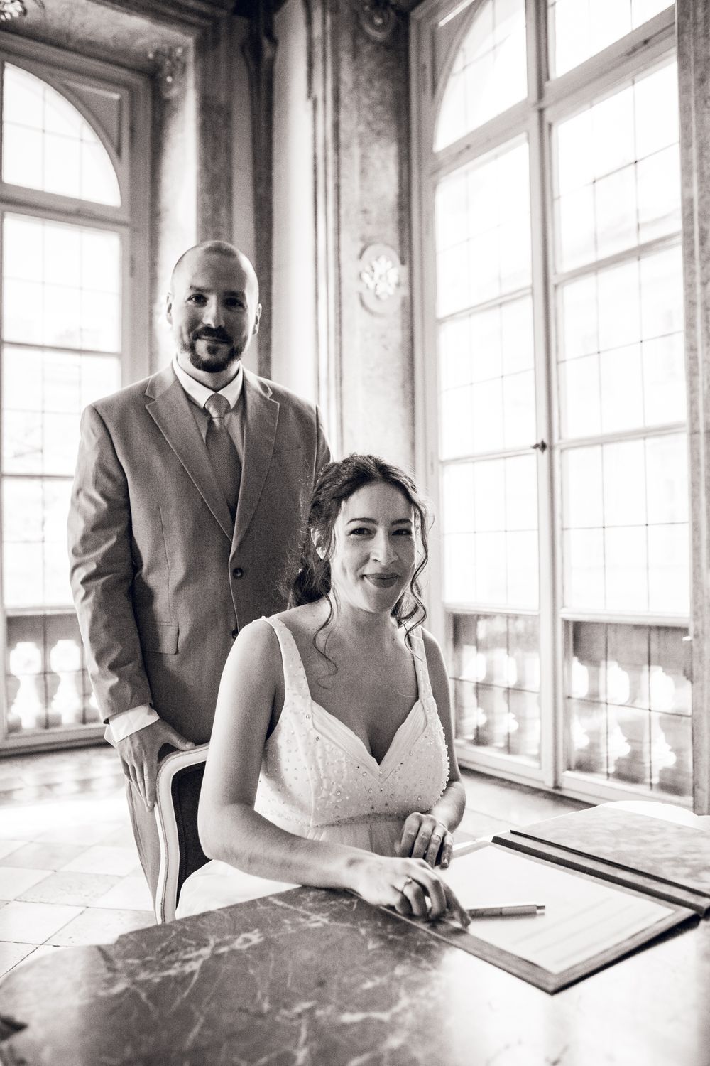 Bride and groom pose at marble table signing wedding register in elegant sunlit room with tall arched windows.