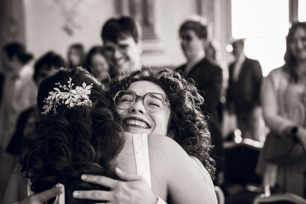 Joyful bride with hair accessories embracing a smiling woman with curly hair and glasses at wedding reception.