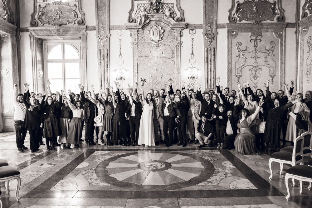 Black and white wedding group photo with guests cheering in an ornate baroque ballroom.